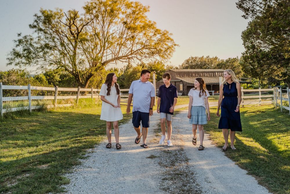 A group of people walk together on a gravel path bordered by white fences during golden hour at a rural property.