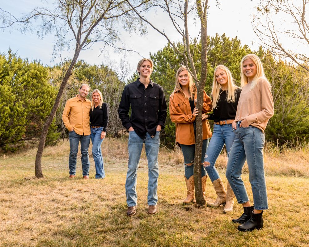 Group of people standing together outdoors in casual fall clothing on grassy terrain with greenery in background.