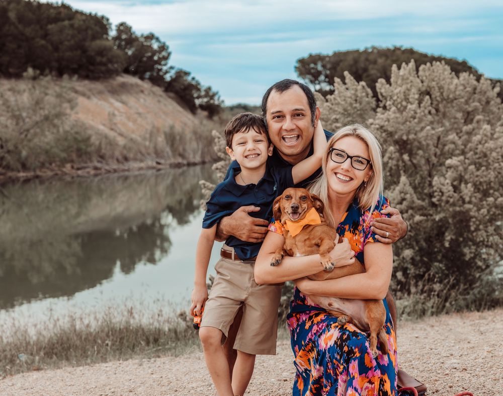 A family posing together with their small fluffy dog near a tranquil lake surrounded by trees.