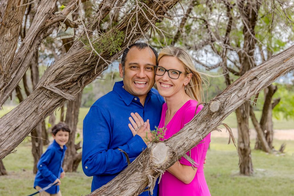 A couple in bright athletic wear smile together by a tree trunk while a child plays in the background.