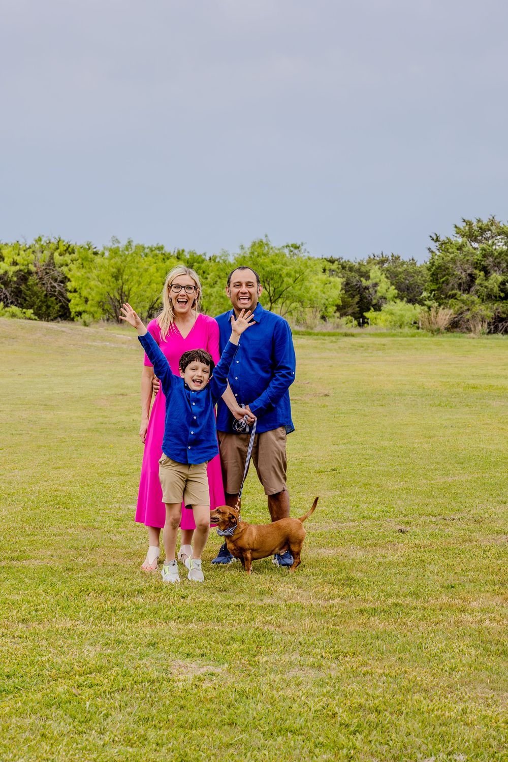 Family poses together on grassy field with their small brown dachshund dog on a sunny day.