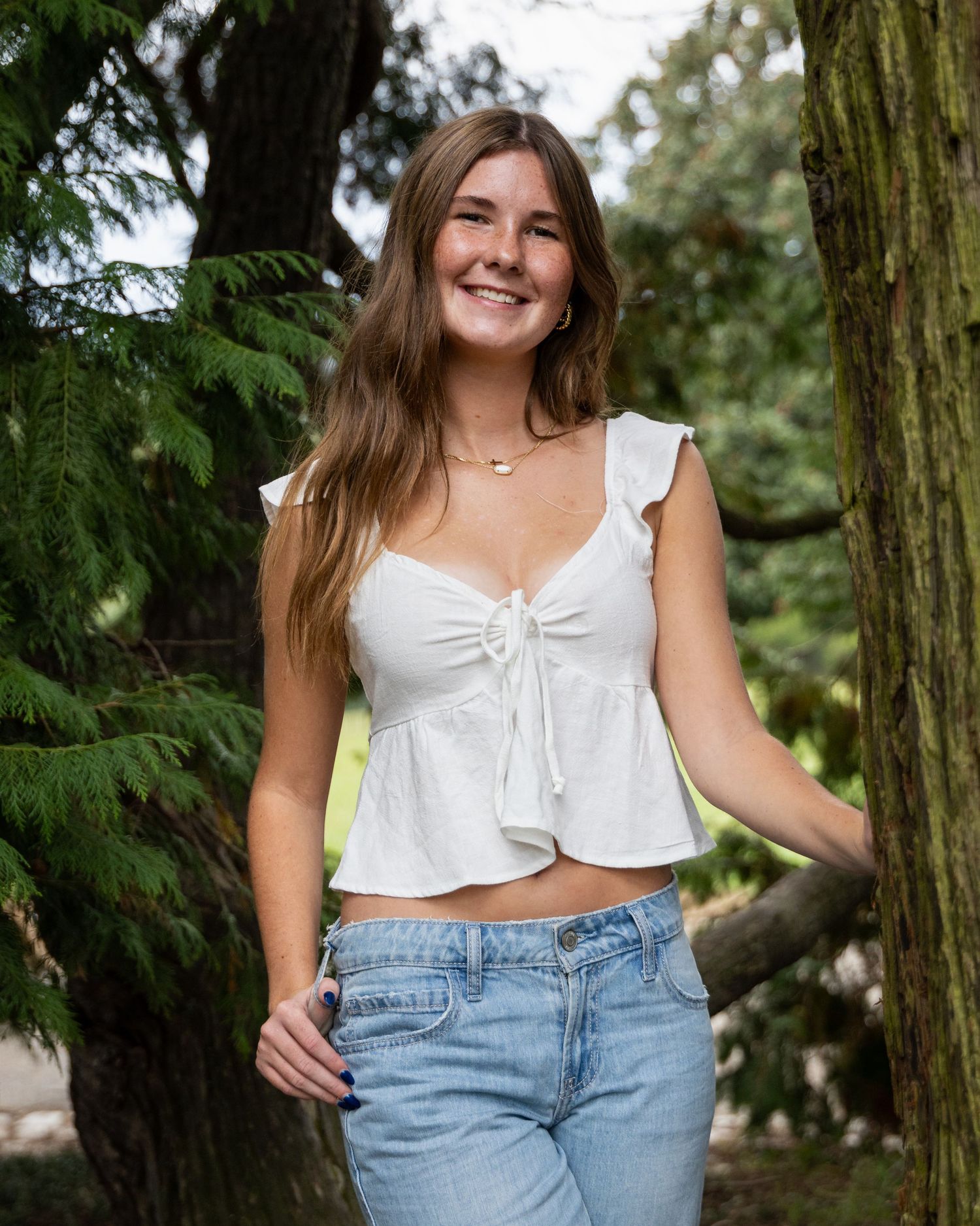Person in white crop top and jeans posing near trees in outdoor photoshoot.