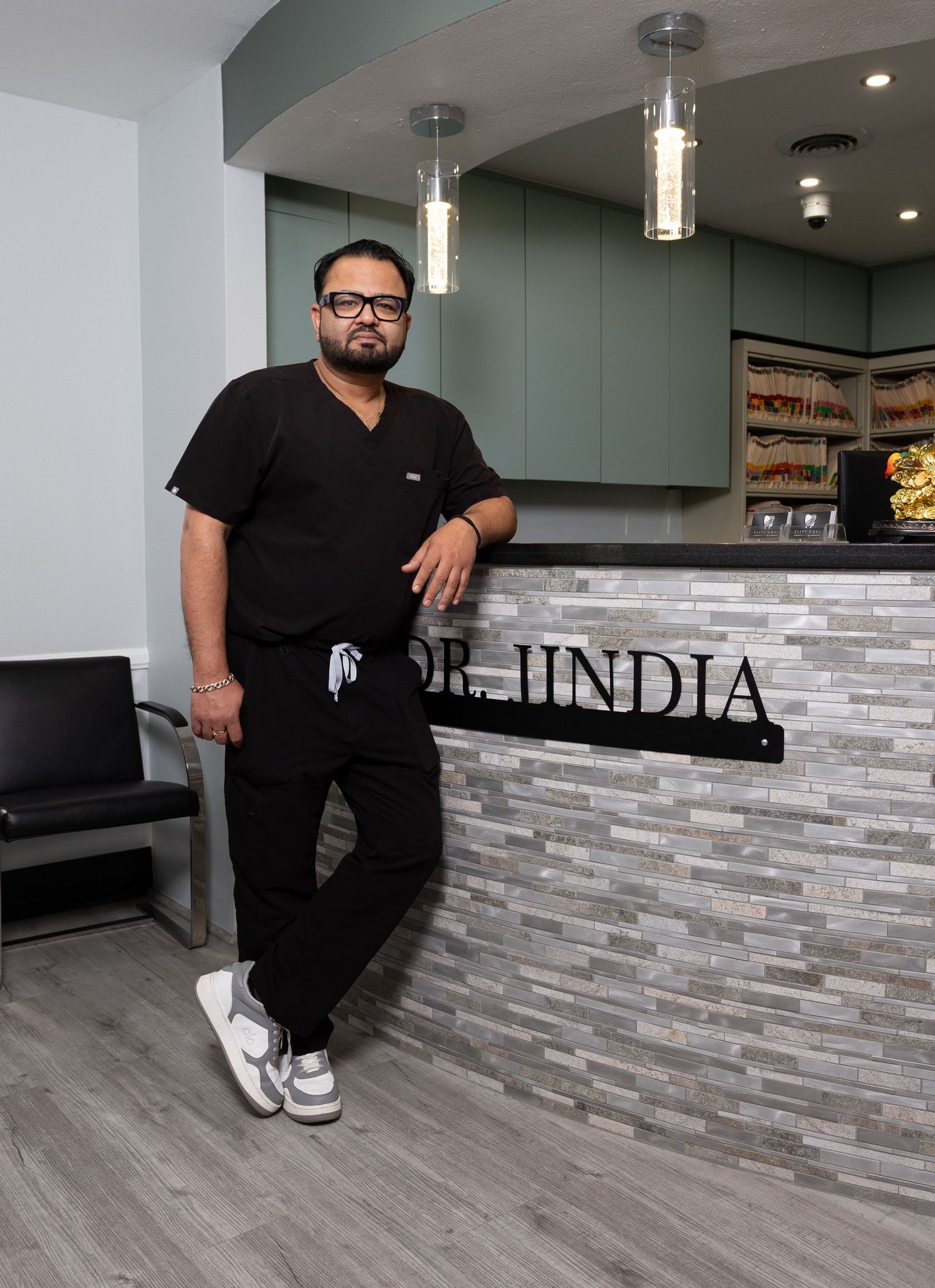 Dr. Jindia poses in black scrubs leaning against a modern stone reception desk in a professional medical office.