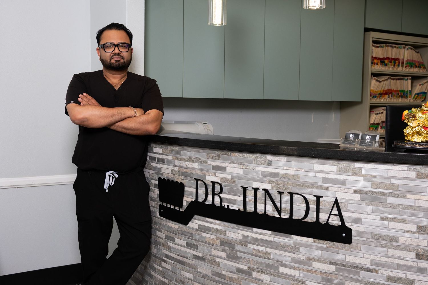 Dr. Jindia poses confidently in black scrubs, arms crossed, beside a modern dental office reception sign.