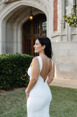 Bride in backless white gown poses outdoors near stone archway, showcasing elegant low back wedding dress design.