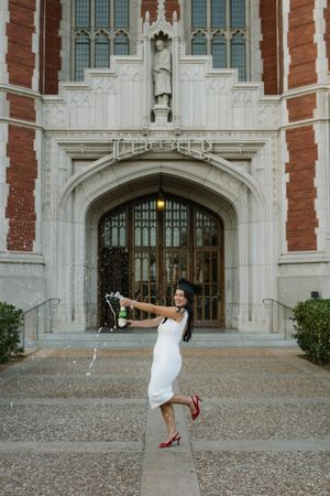 Graduate in white dress and red heels celebrates with champagne outside ornate university building.