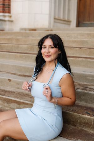 A woman in a light blue dress sits on stone steps holding a stethoscope, smiling for a nursing graduation photo.