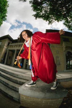 Joyful graduate in red cap and gown poses on brick wall outside school building wearing Converse sneakers.