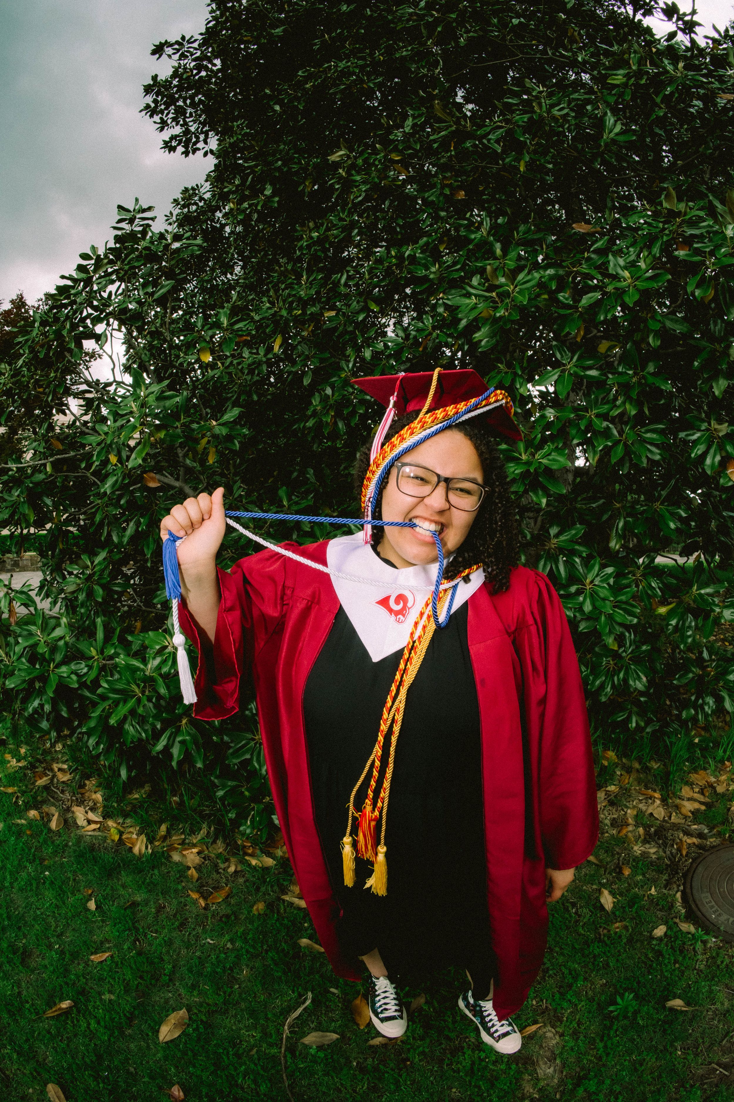 Smiling graduate in red cap and gown holding honor cords outdoors near green trees on graduation day.