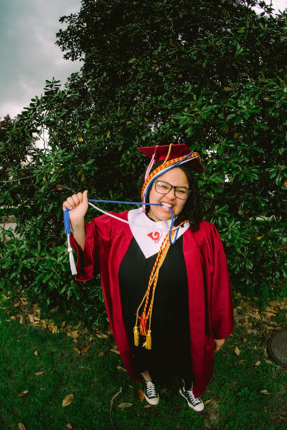 Smiling graduate in red cap and gown holding honor cords outdoors near green trees on graduation day.