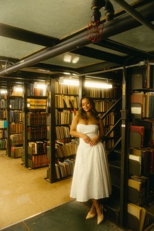 Woman in white strapless dress poses among towering bookshelves in a vintage library setting.