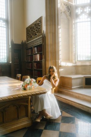 Bride in white dress sitting at wooden table in sunlit gothic library with floral bouquet.