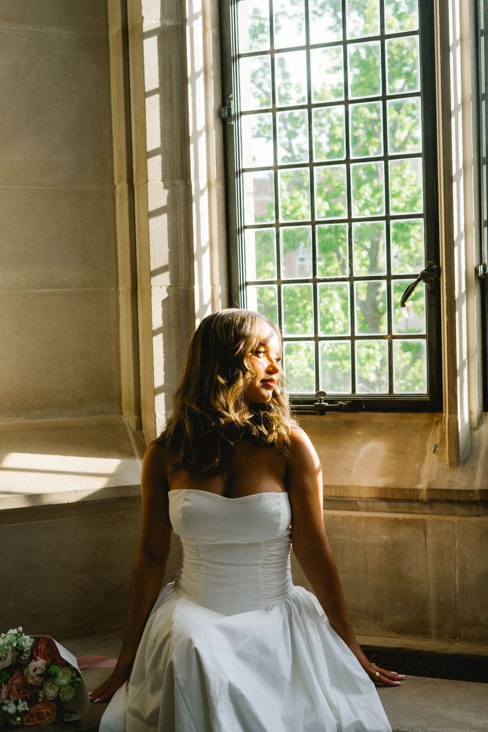 Bride in white strapless gown sitting by a sunlit leaded glass window, gazing thoughtfully to the side.