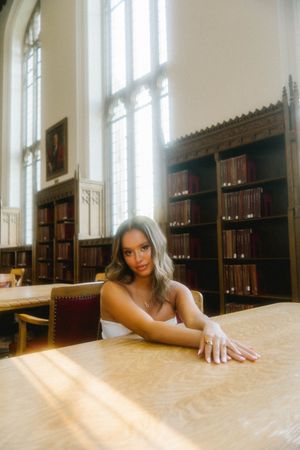 Young woman in white dress poses at wooden table in sunlit classic library with tall windows and bookshelves.