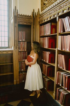Woman in white dress holding red book in ornate wooden library with shelves of red books.