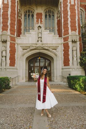 A smiling graduate in white dress and maroon stole holds flowers outside a Gothic brick university building.