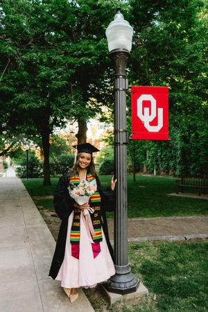 Graduate in cap and gown holds flowers beside an OU banner on University of Oklahoma campus.