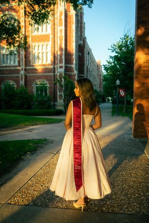 Graduate in white dress with red sash poses on campus walkway beside brick university building.