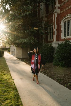 Graduate in cap and gown walks along sunny campus sidewalk beside brick building, wearing red stole and heels.