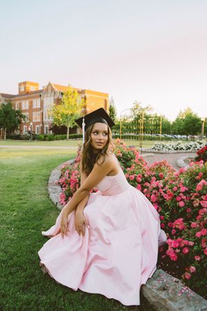 Young woman in pink ball gown and graduation cap poses by pink roses on a college campus at dusk.