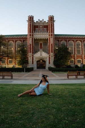 A graduate in cap and white dress poses on grass before a grand red brick university building at dusk.