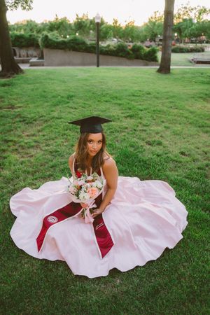 Graduate in pink ball gown and cap sits on green grass holding a floral bouquet during outdoor graduation photo.