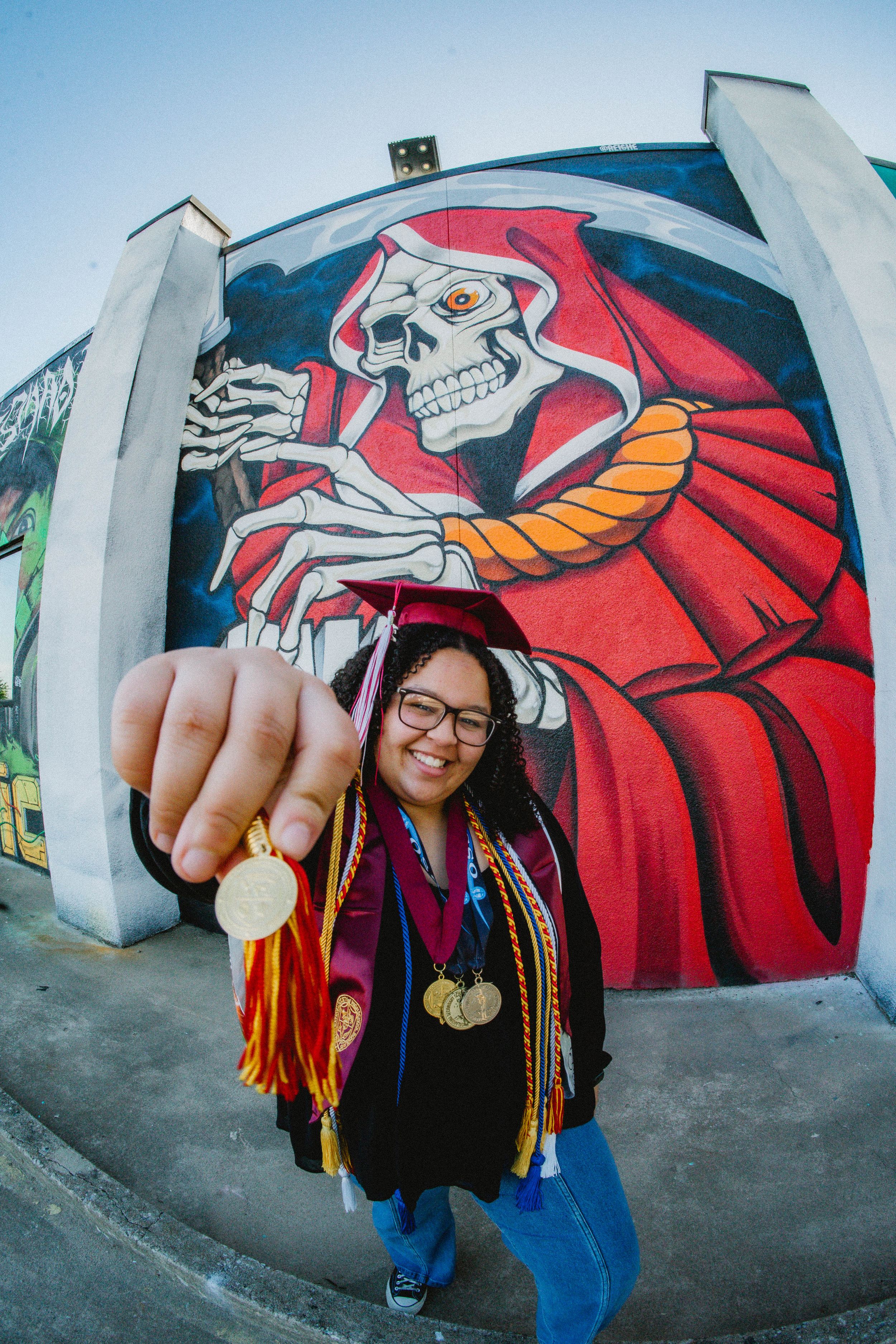 Graduate in cap and gown holds medal toward camera in front of colorful skeleton mural street art.