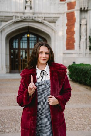 Young woman in red fur coat and houndstooth dress pointing up, standing before ornate stone building entrance.