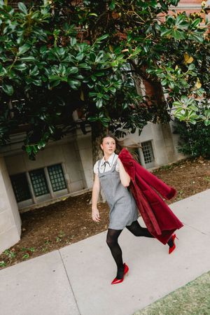 Woman in gray dress, black tights, red heels carries a red coat while walking outdoors near a large green tree.