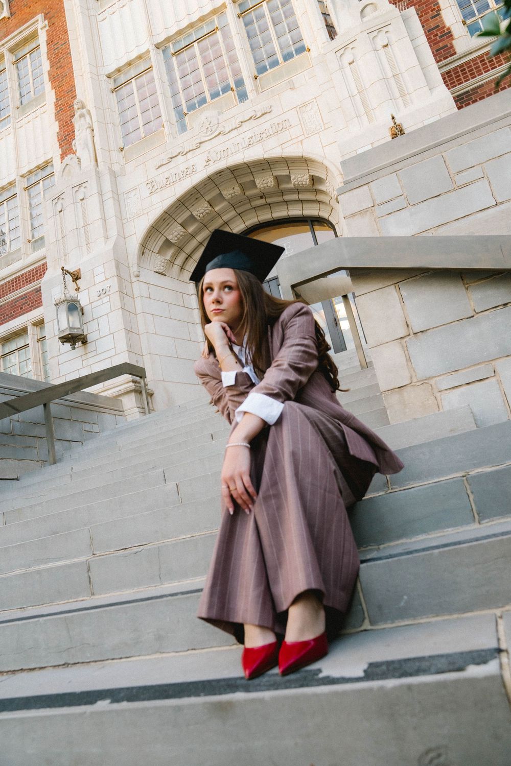 A young woman in graduation cap and brown gown sits on university steps wearing red heels, looking contemplative.