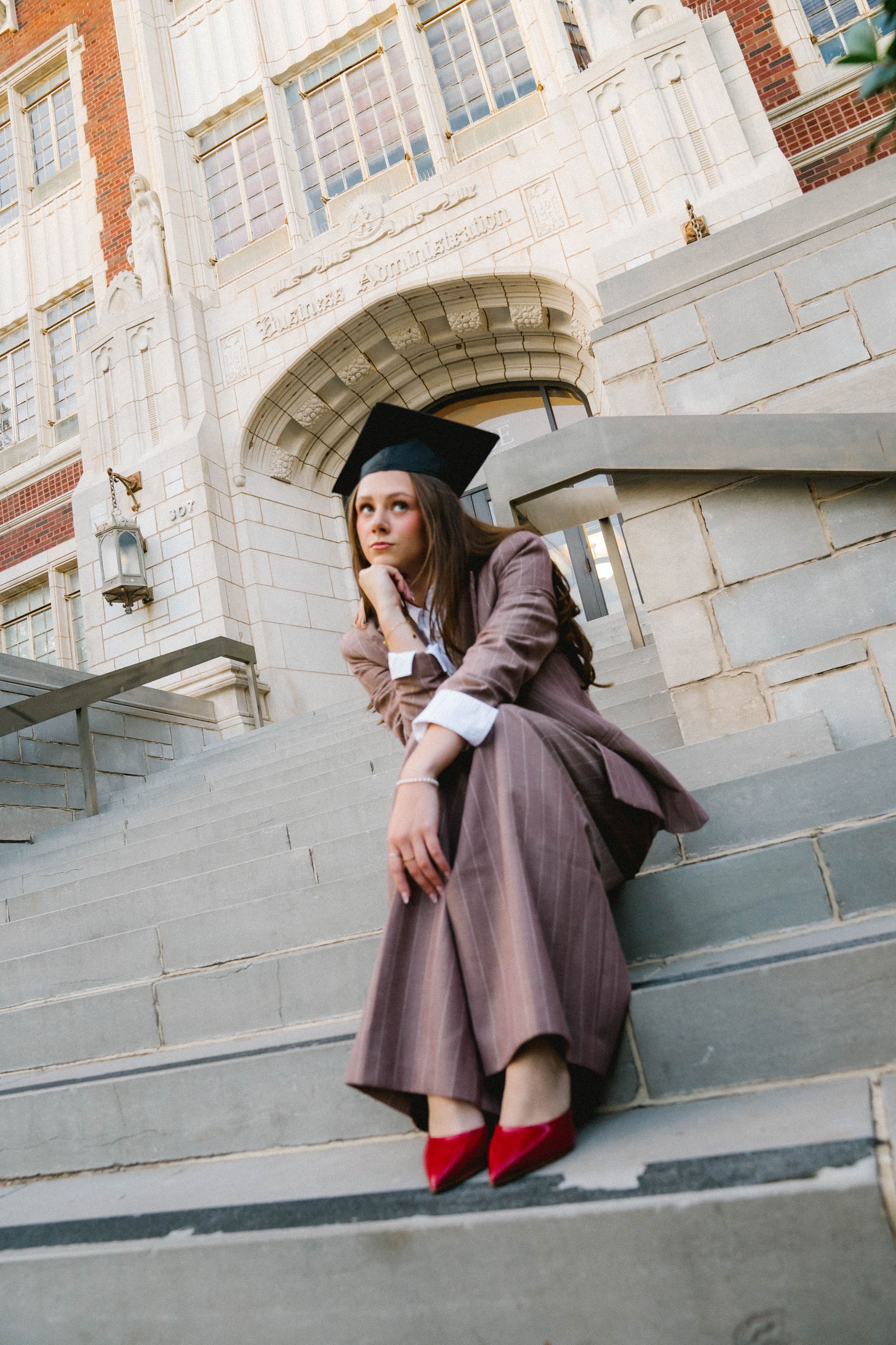 A young woman in graduation cap and brown gown sits on university steps wearing red heels, looking contemplative.