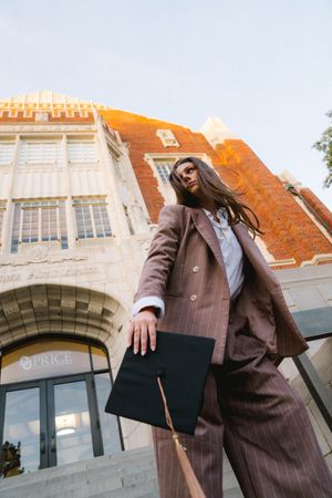 Graduate in brown pinstripe suit holding cap outside USC Price School building.