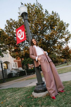 Woman in brown suit posing by a lamp post with University of Oklahoma flag on a college campus.