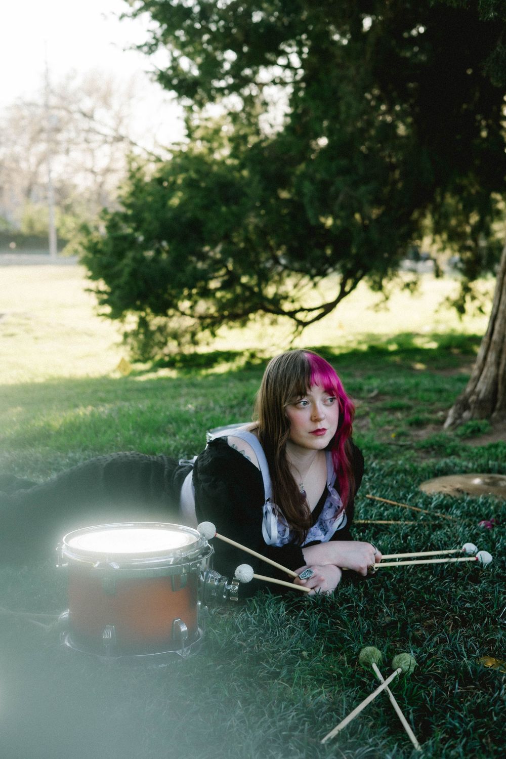 Young woman with pink hair lying on grass in a park with drum sticks and a snare drum beside her.