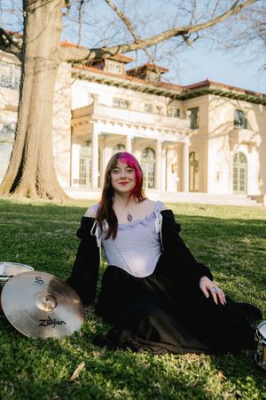 Young woman with pink hair sits on grass with Zildjian cymbal near a white mansion.