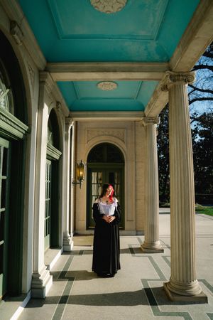 Woman in black skirt and white top poses on elegant neoclassical portico with teal ceiling and marble columns.