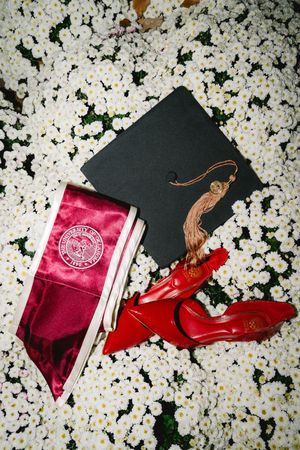 Graduation cap with tassel, red heels, and maroon sash on white flower bed.