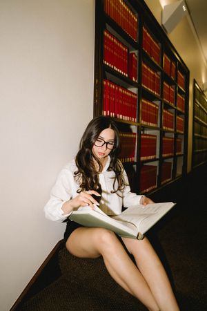 Young woman in glasses reading large book while sitting on floor in law library.