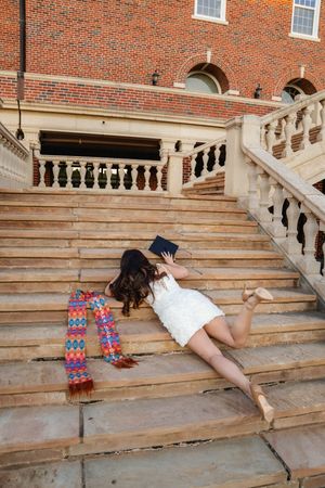 Young woman in white dress lying on brick building steps with colorful scarf and book nearby.
