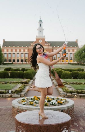 Woman in white dress celebrating graduation, popping champagne in front of a brick university building with manicured gardens.