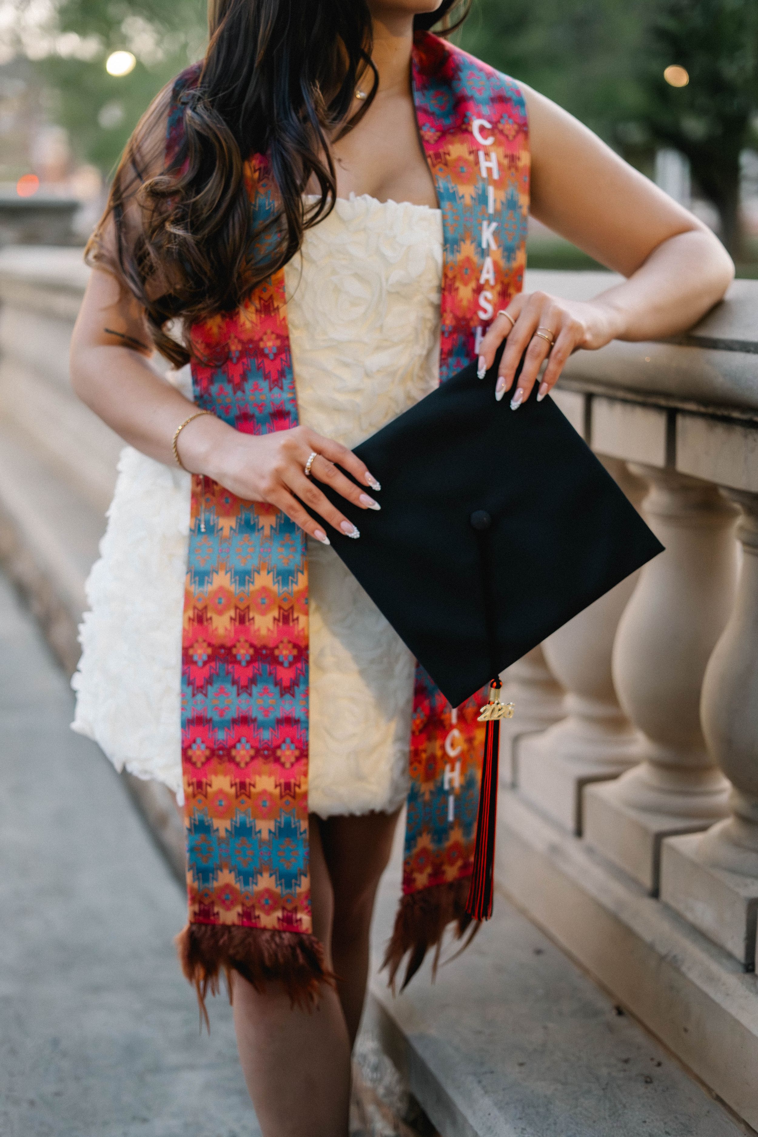 Graduate holding black cap wearing colorful Chickasaw stole over white dress at outdoor ceremony.