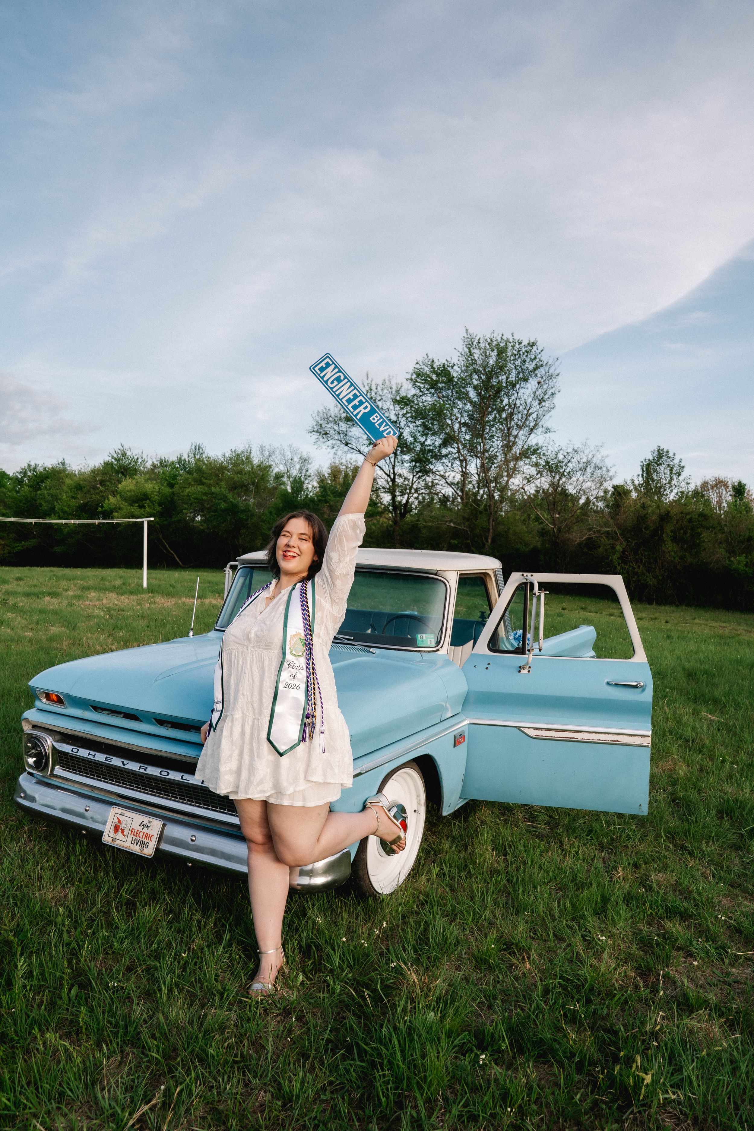 Graduate in white dress holds diploma beside vintage light blue pickup truck in green field.