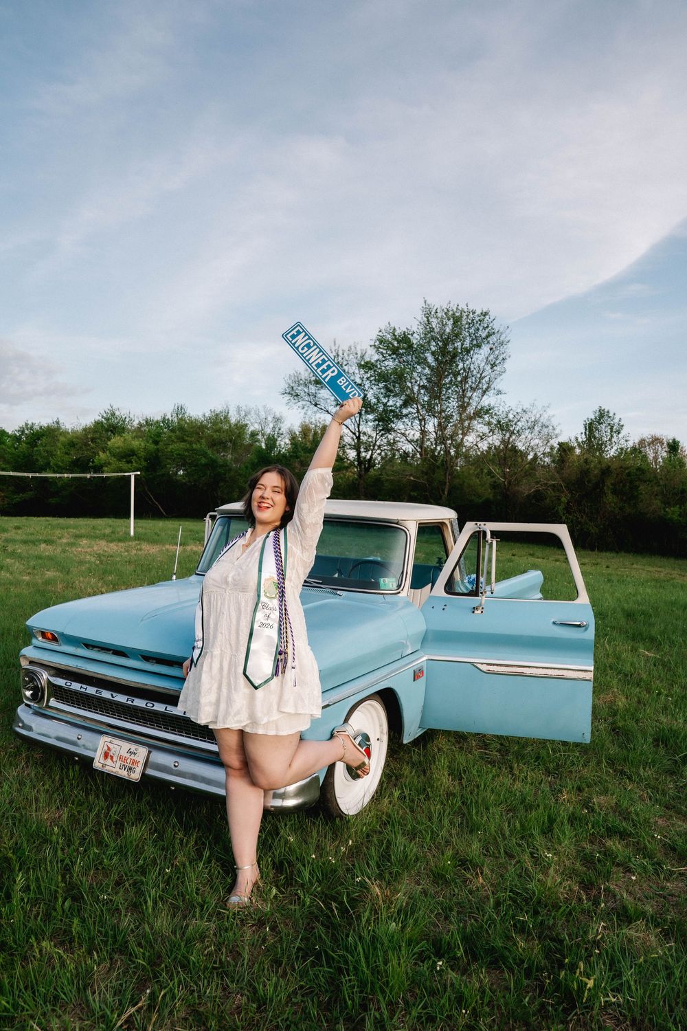 Graduate in white dress holds diploma beside vintage light blue pickup truck in green field.