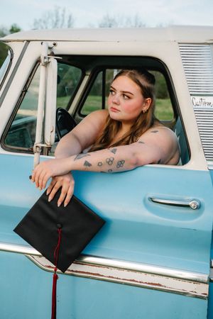 Young woman with tattoos leaning out of a vintage blue truck window, holding a black graduation cap with red tassel.