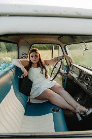 Young woman in white dress poses in vintage teal and white truck cab, leaning on steering wheel in a field.