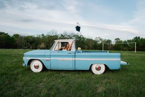 A vintage light blue pickup truck parked on green grass with a graduation cap tossed in the air.