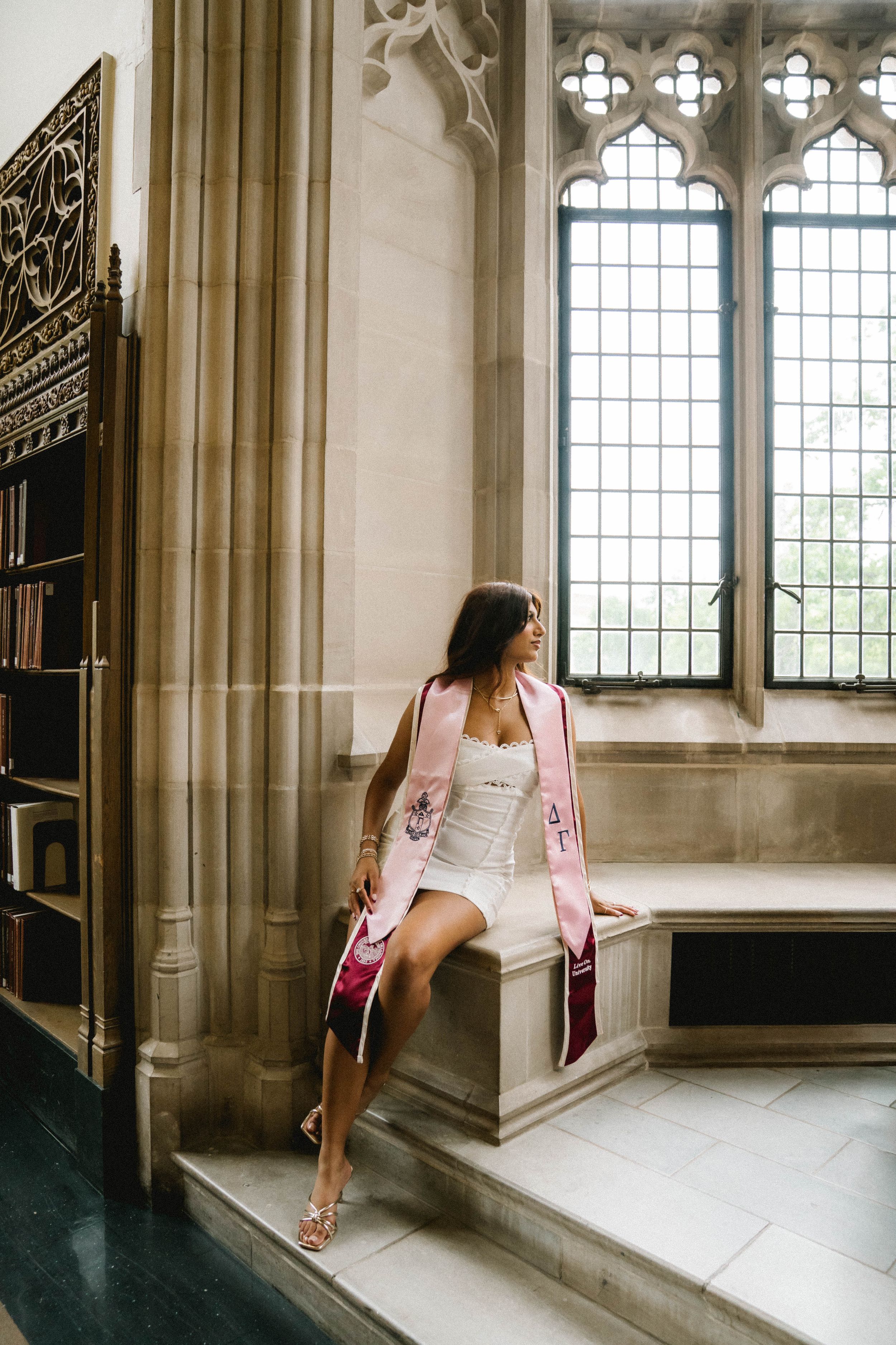 A woman in a white dress and pink graduation stole sits on a stone ledge in an elegant gothic library.