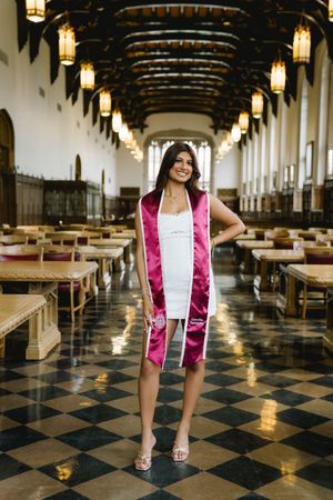 A graduate poses in a white dress with a pink satin stole in an elegant hall with checkered floors and hanging lanterns.