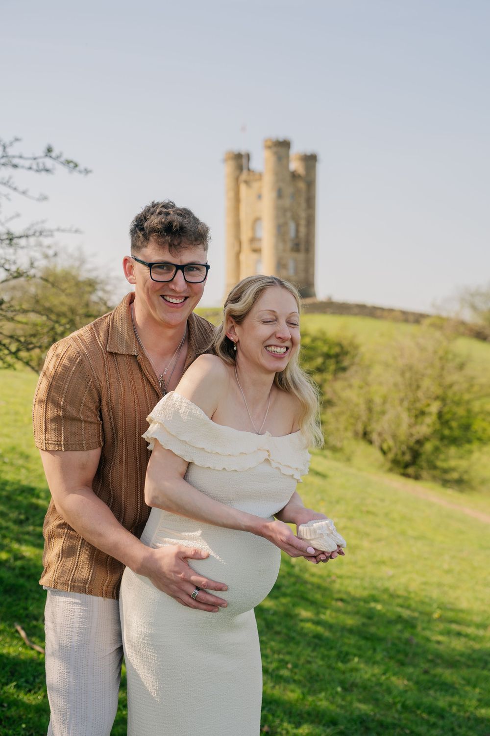 Happy couple posing outdoors near a historic tower, with the woman in a white dress showing her baby bump.