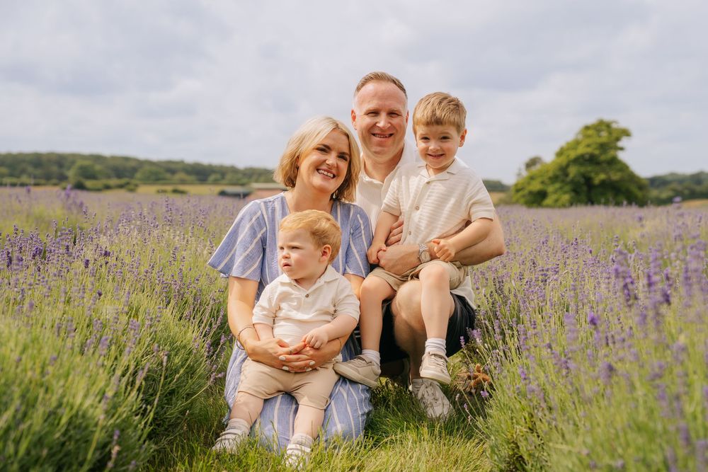 Family of four smiling in a lavender field, parents holding two young boys surrounded by purple lavender fields.